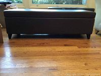 Side view of a dark brown leather bench ottoman with short legs on a wood floor.