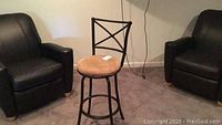 Bar stool positioned between two black chairs, showing the metal frame, cross-back design and stained suede seat cushion.