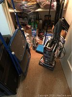 View of blue metal step stool, Shark vacuum cleaner, and various cleaning supplies and bags in the background showing contents of lot.
