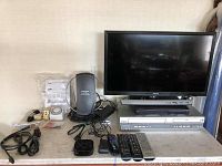 Front view of Samsung 23-inch LED TV, Panasonic DVD/VHS player underneath, Samsung Blu-ray player, Roku device, remote controls, power adapters, and cables spread on a table.