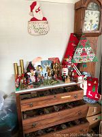 Top view on dresser with various vintage Christmas figurine ornaments including angels, Santa Claus, and assorted characters, along with a quilted Christmas tree decoration standing next to the clock. Cloth stockings draped over the side of the furniture.