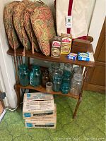 Photo showing wood table with shelf, 23 glass canning jars in clear and blue bubble glass on shelf, fabric chair pads stacked on top