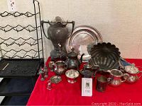 Photo of silverplate items on red cloth including serving plate, coffee urn, sugar and creamer, salt and pepper, teapot, fluted molds, and other smaller pieces, next to wire wine rack