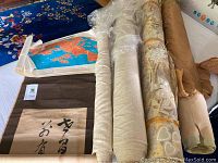 Image shows four bolts of upholstery fabric in beige tones with different textures and patterns, a rolled Japanese scroll with Asian calligraphy and a bright colored map, plus a collection of rolled maps.