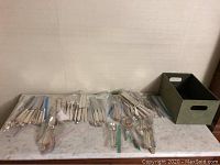 Overview of all silver plated cutlery grouped in plastic bags on table surface next to storage box.