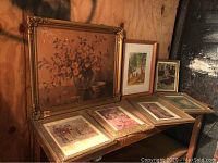 Wide view of six framed art pieces arranged on a wooden bench and leaning against a wall. Large floral painting is prominent with visible wear and frame damage. Several smaller prints and a watercolor painting are laid out beneath and beside it.
