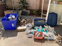 Overview of lot showing blue bins with various hardware parts, metal work pedestal with wheels, and assorted components on concrete surface.