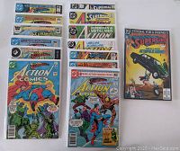 14 Action Comics and Superman comic books laid out showing covers with various characters including Superman, Supergirl, and villains. Covers show colorful illustrated scenes with varying wear.