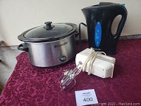 Photo showing three home kitchen appliances: Bravetti crockpot, electric kettle, and handheld mixer on a table.