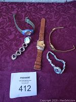 Five assorted women's watches laid out on a purple patterned fabric, including a brown leather Winnie the Pooh watch, silver metal linked watches, a blue plastic watch, and a gold-tone bangle watch. A MaxSold lot number tag 412 visible in lower left.
