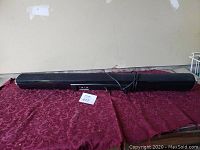 Black wireless sound bar speaker placed on a red patterned cloth against a plain wall, shows front panel with control buttons and connected secondary component.