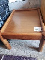 Hardwood side table in light colour, showing top surface and part of legs with raised edges, placed on concrete floor next to a black container.