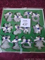 Box containing 12 crystal Christmas ornaments with white bows and blue roses, arranged in a green tray. Ornaments in various shapes including angels, bells, stars, Christmas trees, and wreaths, each with a hanging string.