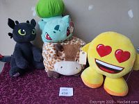 Four large stuffed animals placed on a maroon patterned fabric background: Night Fury dragon Toothless plush, Bulbasaur Pokemon plush with tag, plush giraffe, yellow heart-eyes love emoji plush with tag.