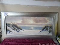 Framed beach landscape painting showing sand dunes, sea, sailboats, and sky.