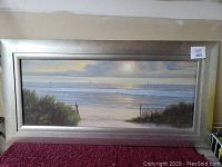 Front view of framed beach picture showing the beach scene with sand, plants, fencing, ocean waves, and sailboats on horizon.