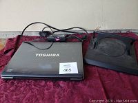 Photo showing Toshiba laptop closed on left, black laptop cooling stand on right, and laptop power adapter cable above them on a red fabric surface.