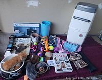 Photo of dog and cat accessories showing paw cleaner, pet bowls, dog mesh gate, collars, bandanas, plush and rubber toys, and books on pet care.