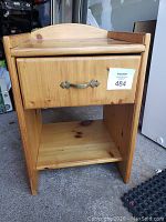 Photo shows light colored wooden night stand with one drawer featuring a decorative metal handle and open shelf below. Slight wear visible on wood surface.