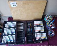 Wooden art easel laid flat on ground with open paint kit showing multiple paint tubes, colored pencils, oil pastels, and three cups filled with colorful markers.