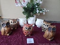 Image of ten owl-themed decorative items on a burgundy cloth, including six brown owl figurines, one red owl figurine, and three white owl ceramic vases or containers, arranged in two rows.