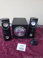 Photo showing two satellite speakers, a central subwoofer with a large silver knob, and a separate wired control knob on a purple patterned cloth.