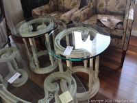 Pair of carved wood end tables with clear round glass tops on hardwood floor, next to fabric armchairs.