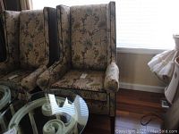 Pair of matching wing chairs with floral upholstery and velvet sides next to a window with light filtering through.