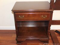 Front view of one Ethan Allen nightstand showing single drawer with brass tone handles, open storage area behind double doors, and carved wooden feet on hardwood floor.