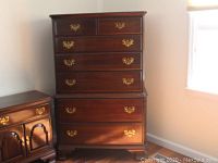 Photo showing front view of tall chest over chest dresser with brass-tone drawer pulls, wood finish, and some reflecting sunlight.