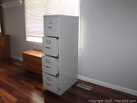 Full side angle view of the gray metal four-drawer filing cabinet placed on hardwood floor.