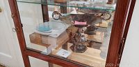 View of the antique silver plate centerpiece bowl with sculpted base, Dansk salt and pepper shakers, and Princess House plates in wooden display cabinet.