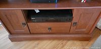Close-up of wooden corner TV cabinet with VHS player on middle shelf, showing metal ring handles and drawer below.