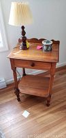 Antique wooden side table with drawer, toleware box, and lamp shown on wooden floor near window.
