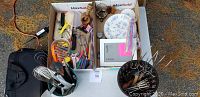 Top-down photo showing contents of boxes and containers on white table: plates, utensils, glassware, and miscellaneous items including pencils and ice scraper.