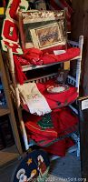 White wicker three-tier shelf holding multiple Christmas-themed linens including placemats, runners, tablecloths and a framed cross-stitch artwork.