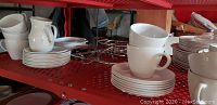 Shelving with stacked white CorningWare cups, saucers, plates, Correlle carafe, warming trivets