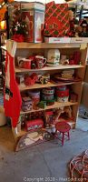 Photo showing wooden shelf stocked with various Christmas-themed mugs, tins, boxes, and ornaments. A red oven mitt hangs on the side, and a small red stool is positioned near the bottom shelf along with some holiday tins and trays.
