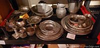 Shelf holding an assortment of pewter tableware pieces including teapots, pitchers, trays, plates, and cups arranged in a cluttered manner.