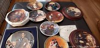 Collection of Norman Rockwell collectible plates laid out on table, showing various scenes and some plates stacked.