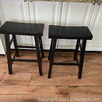 Front view of two black wooden counter stools with saddle-shaped seats on a wood floor in front of white cabinetry.