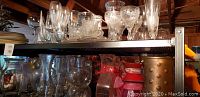 Two shelves of assorted glassware including a punch bowl, beer glasses, wine glasses and multiple vases, displayed on a black metal shelving unit under wooden ceiling beams.