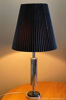 Front view of metal table lamp with black pleated shade lit up on wooden surface.