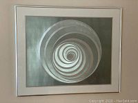 Full view of the framed wall art showing the concentric spiral design in metallic silver and green tones with white border and silver frame.