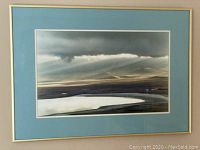 Framed artwork showing a wide view of Ngorongoro Crater with lake, hills, and cloudy sky. Blue mat and gold frame visible.