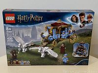 Front view of the LEGO Harry Potter set box showing the Beauxbatons Carriage and winged horses, with minifigures and Hogwarts castle backdrop.