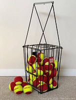 Wilson Starter Red red and yellow felt junior tennis balls inside and outside of the black metal Wilson ball hopper with handle, placed on carpet flooring against a white wall.