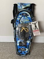Front view of a pair of Yukon Charlie's blue/gray camo junior youth snowshoes with bindings and yellow laces, showing included carry bag and poles in packaging.