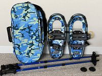 Complete set showing two junior snowshoes, two blue adjustable ski poles, and matching camo carry bag on a floor against wall.