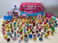 Wide view showing the pink and blue Ice Cream Truck playset surrounded by numerous colorful Shopkins figurines in various forms, including food items and household objects, along with a pink shopping basket and blue shopping cart.